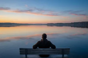 Person in olive jacket sitting alone on weathered wooden bench facing tranquil lake at sunset, with peach and blue clouds reflected in still water and dark forested shoreline in distance