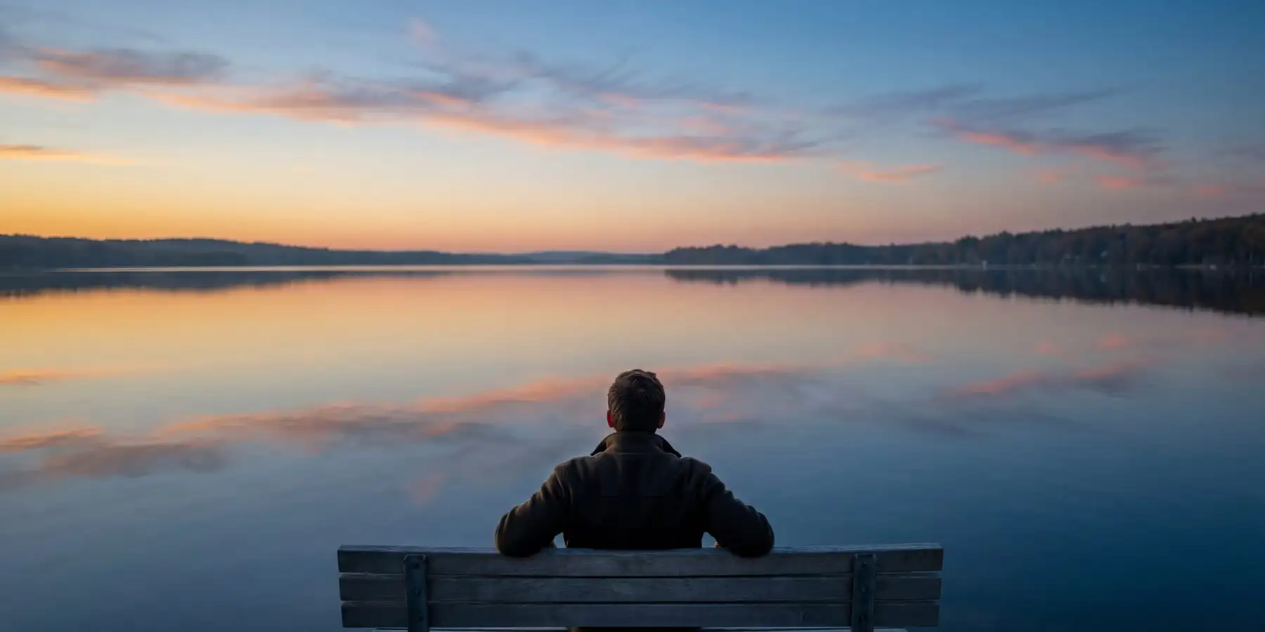 Person in olive jacket sitting alone on weathered wooden bench facing tranquil lake at sunset, with peach and blue clouds reflected in still water and dark forested shoreline in distance
