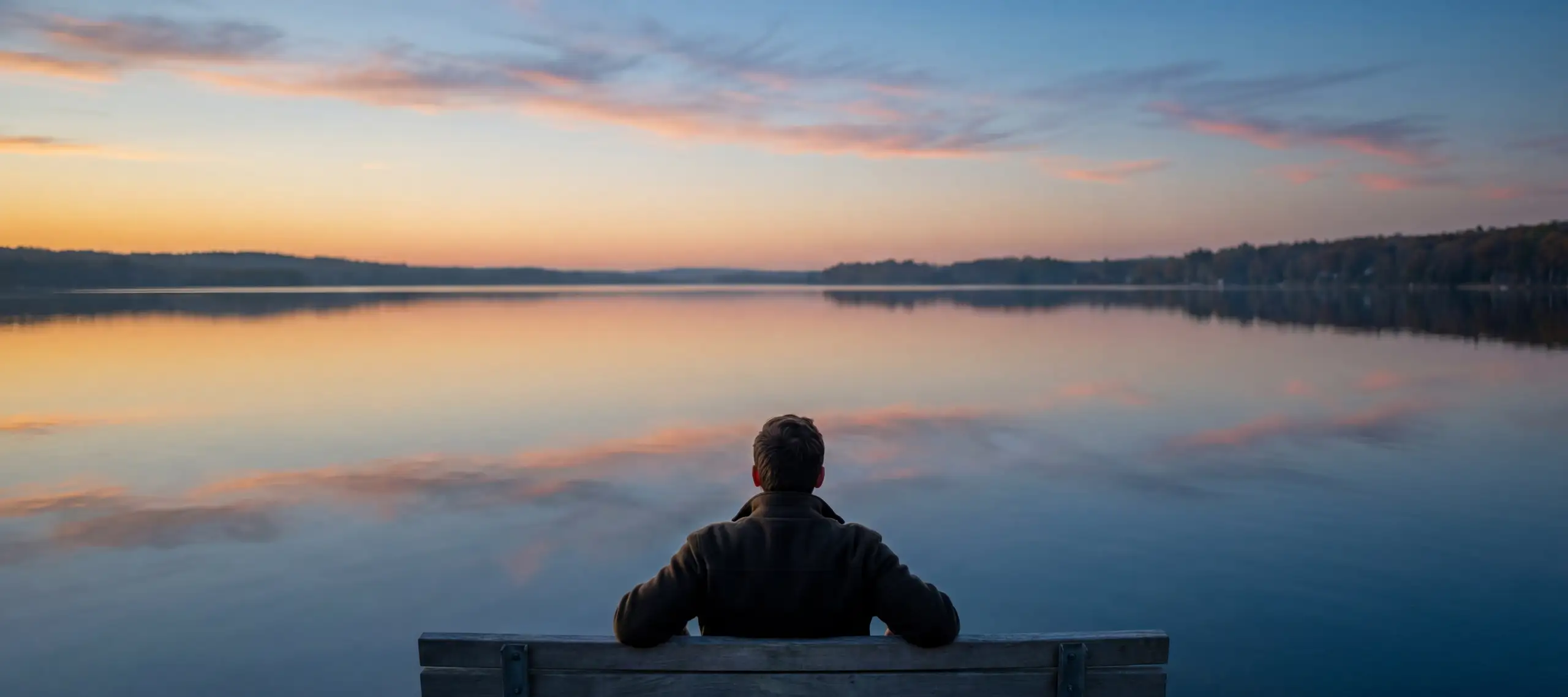 Person in olive jacket sitting alone on weathered wooden bench facing tranquil lake at sunset, with peach and blue clouds reflected in still water and dark forested shoreline in distance