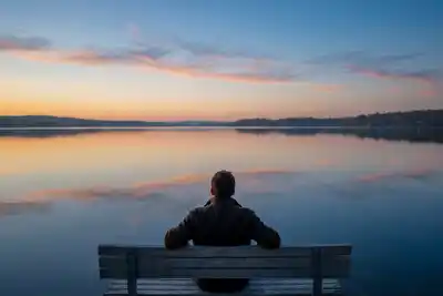 Person in olive jacket sitting alone on weathered wooden bench facing tranquil lake at sunset, with peach and blue clouds reflected in still water and dark forested shoreline in distance