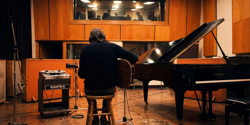 A musician with shoulder-length hair sits on a wooden stool in a warm-toned recording studio, holding an acoustic guitar, facing a black Bechstein grand piano with its lid raised, while producers watch from a lit control room window above