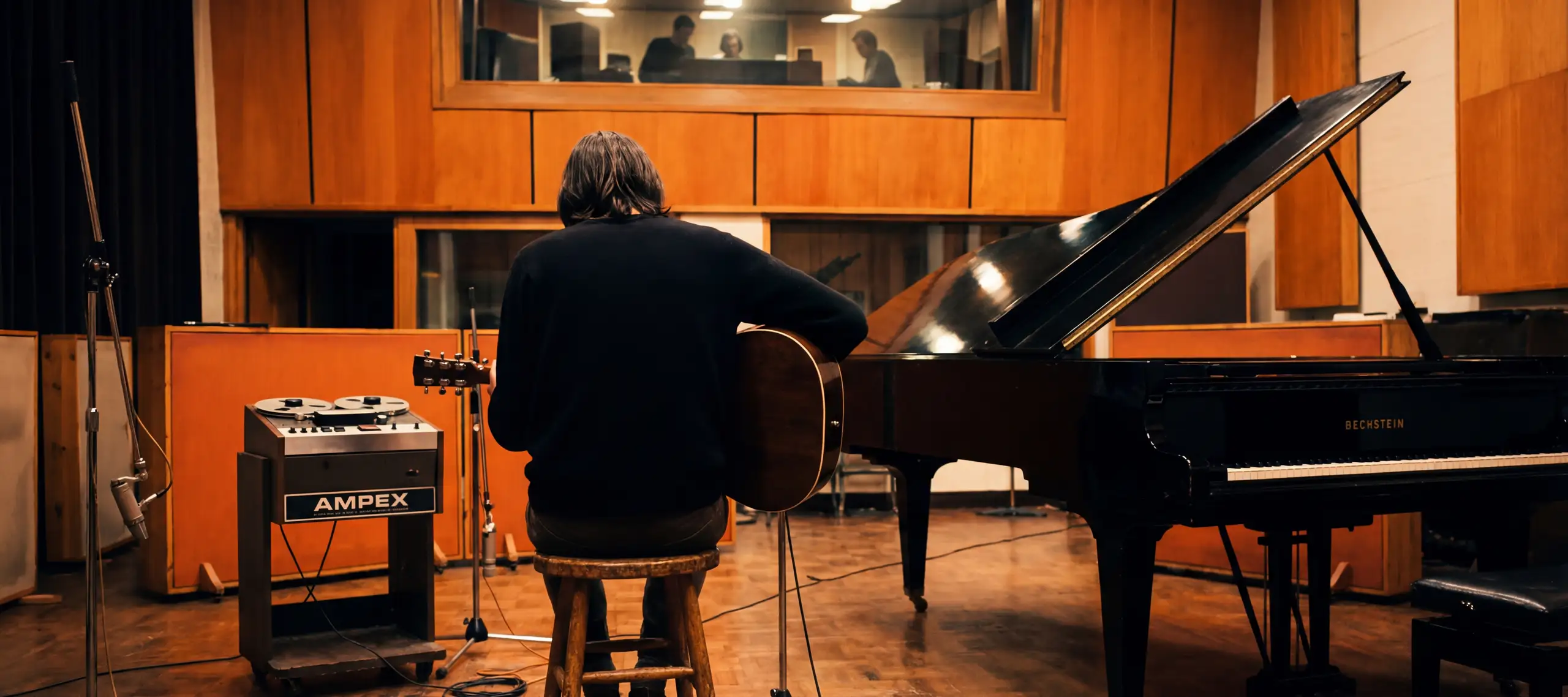 A musician with shoulder-length hair sits on a wooden stool in a warm-toned recording studio, holding an acoustic guitar, facing a black Bechstein grand piano with its lid raised, while producers watch from a lit control room window above