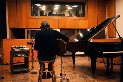 A musician with shoulder-length hair sits on a wooden stool in a warm-toned recording studio, holding an acoustic guitar, facing a black Bechstein grand piano with its lid raised, while producers watch from a lit control room window above